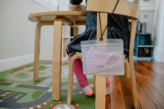 Image of a child, sitting on a wooden chair at a desk in a children's playroom. There is a face mask hanging off the back of the chair in a plastic case with the words 'Atoms everyday face mask' printed on it.