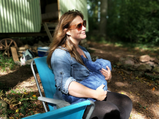 Image of a woman seated on a camping chair breastfeeding a baby at a campsite